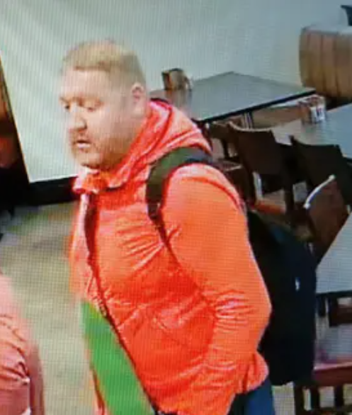 Man in orange jacket with a black backpack who has light hair and a short beard.