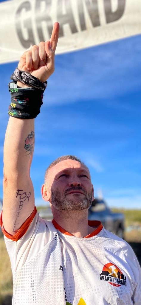 Gary McKee a white man with grey hair and a short beard, wearing a white t shirt, pointing his finger in the air with the words 'for Bill' written on his extended arm.